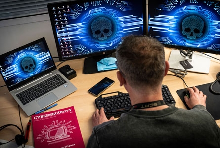 Une personne devant des écrans avec une tête de mort et une documentation sur la cybersécurité.