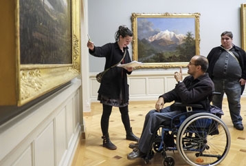 Une personne en chaise roulante au musée devant un tableau avec un guide
