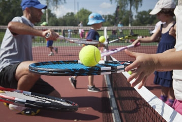enfants tenant leur raquette de tennis au-dessus d'un filet. Un homme accroupi leur explique comment faire
