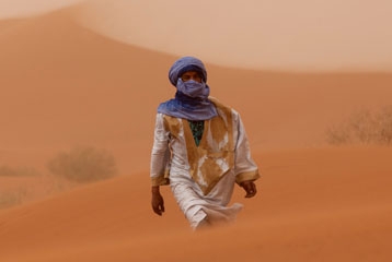 Homme en tunique blanche couverte de sable, coiffé d'un turban bleu, marchant fans des dunes