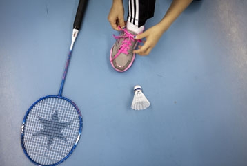gros plan sur une chaussure de sport avec des mains qui refont les lacets roses. A côté, une raquette de badminton et un volant