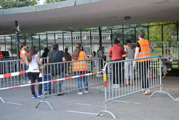 personnes entre barrières faisant la queue, entourées de personnes vêtues d'un gilet orange fluo