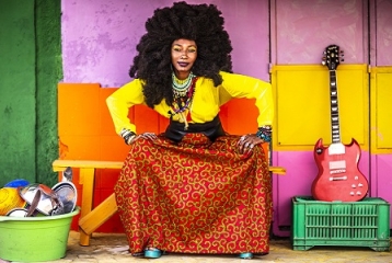 femme à la coupe afro qui se tient assise les mains sur les genoux. Une guitarre électrique est posée à côté